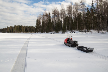 snowmobile with a trailer is on the lake, around the snowy forest in sunny weatherの写真素材