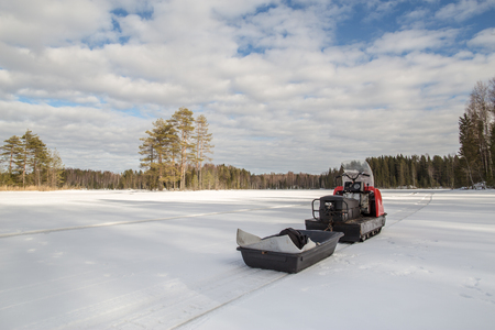 snowmobile with a trailer is on the lake, around the snowy forest in sunny weatherの写真素材