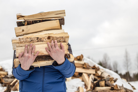 a man carries in his hand a great bunch of chopped firewood in the winter in the village against the backdrop of snow-covered heaps of other woodの写真素材