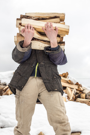a man carries in his hand a great bunch of chopped firewood in the winter in the village against the backdrop of snow-covered heaps of other woodの写真素材
