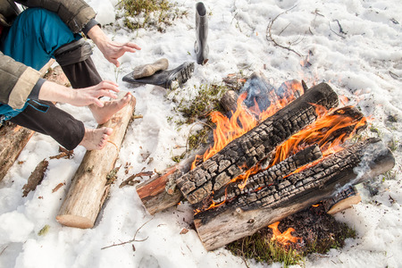 A man warms his hands and feet near the fire in the pine forest covered with snow in winterの写真素材