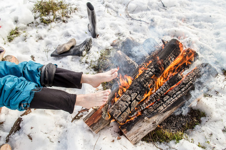 A man warms his barefoot feet near the fire in the pine forest covered with snow in winterの写真素材