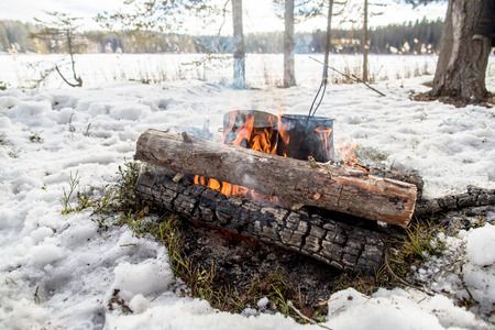 Cooking in a winter hike in the cauldron hanging over the fire in the snow-covered pine forest while camping, from the boiler coming vaporの写真素材