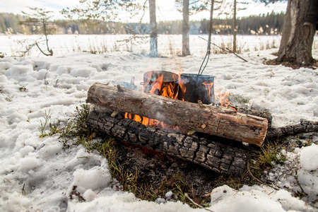 Cooking in a winter hike in the cauldron hanging over the fire in the snow-covered pine forest while camping, from the boiler coming vaporの写真素材