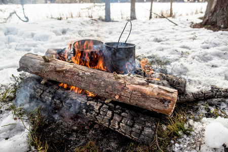 Cooking in a winter hike in the cauldron hanging over the fire in the snow-covered pine forest while camping, from the boiler coming vaporの写真素材