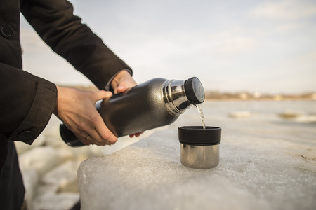Man pours hot tea from a flask into a cup which stands on an ice floe on the river bank in the spring sunny dayの写真素材