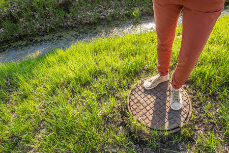 A person standing on of an old rusty manhole cover near gutter with water and algae among fresh spring grass in the parkの写真素材