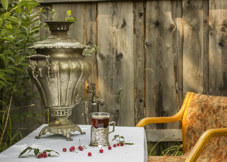 vintage copper samovar in a cup holder and a glass of hot tea stands on the table are scattered cherries and raspberries, as well is worth chair in Russian village outdoorsの写真素材