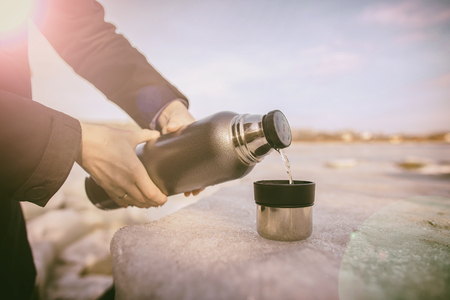Man pours hot tea from a thermos into a cup which stands on an ice floe on the river bank in the spring sunny dayの写真素材