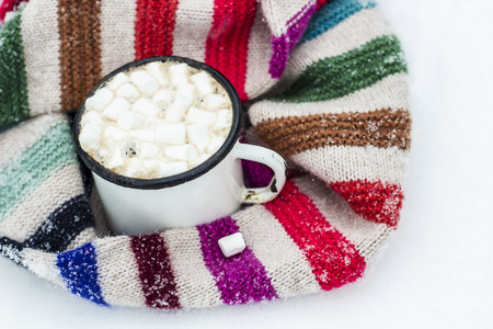 Old metal cup of hot cocoa with marshmallows and a soft colorful knitted scarf on a fluffy the snow close-upの写真素材