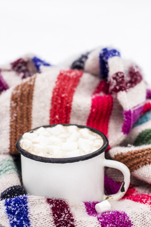 Old metal cup of hot cocoa with marshmallows and a soft colorful knitted scarf on a fluffy the snow close-upの写真素材