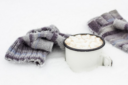 Old metal cup of hot cocoa with marshmallows standing in the snow next to lie a striped knitted the glovesの写真素材