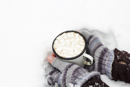Hands adult human in a striped knitted gloves holding an old metal a cup of warm cocoa with marshmallows in a fluffy the snowの写真素材