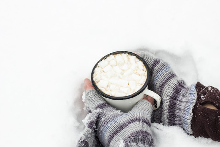 Hands adult human in a striped knitted gloves holding an old metal a cup of warm cocoa with marshmallows in a fluffy the snowの写真素材