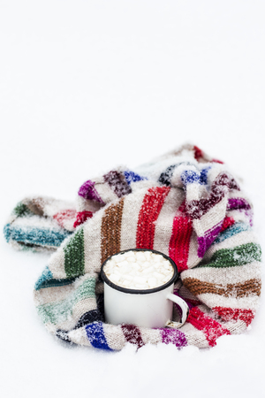 Old metal cup of hot cocoa with marshmallows and a soft colorful knitted scarf on a fluffy the snowの写真素材