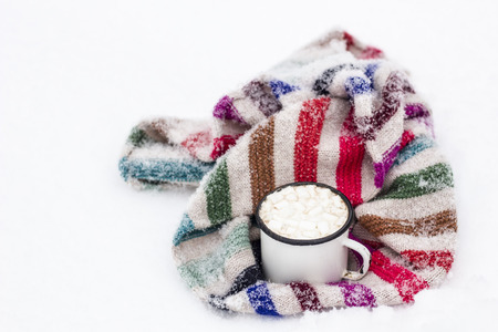 Old metal cup of hot cocoa with marshmallows and a soft colorful knitted scarf on a fluffy the snowの写真素材