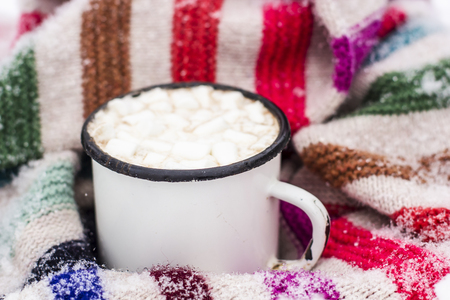 Old metal cup of hot cocoa with marshmallows and a soft colorful knitted scarf on a fluffy the snow close-upの写真素材