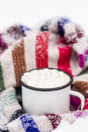 Old metal cup of hot cocoa with marshmallows and a soft colorful knitted scarf on a fluffy the snow close-upの写真素材