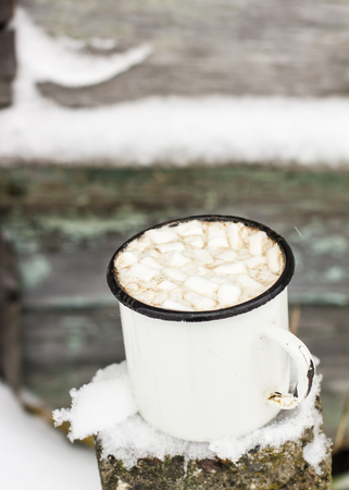 Old metal cup of hot cocoa with marshmallows stands on covered with snow pillar on the background shabby wood wall outdoorsの写真素材