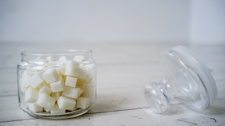 White sugar lumps in a glass jar on the wooden table painted in white color lies next the cover, close upの写真素材
