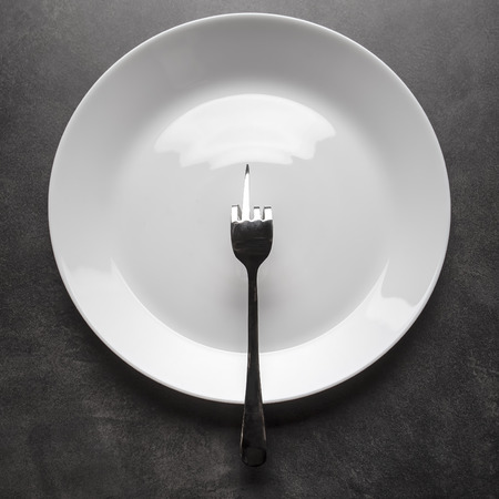 Fork bent in the shape of obscene gesture middle finger sign lies on an empty plate instead of foodの写真素材