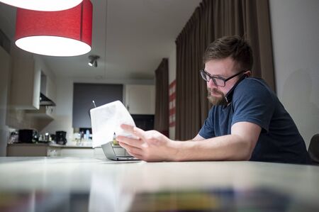 Calm man manages finances by phone while sitting at home in the kitchen, he looks at the receipt he holds in his hand, next to him is an open laptop.の写真素材