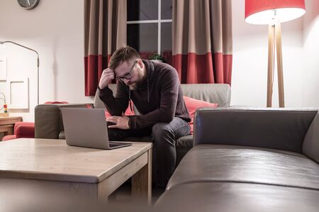 Frowned man looks at his laptop, he reads messages or watches a video that upset him while sitting on a leather sofa at home.の写真素材