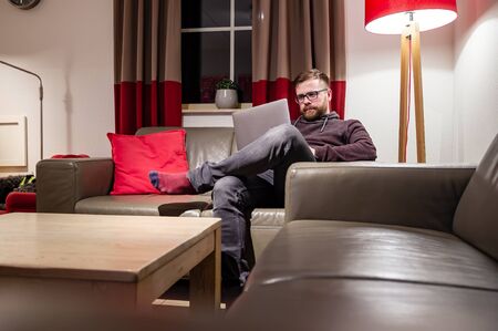 Man looks at his laptop, he reads messages or works, sitting in a comfortable position on a leather sofa, in a cozy home environment.の写真素材