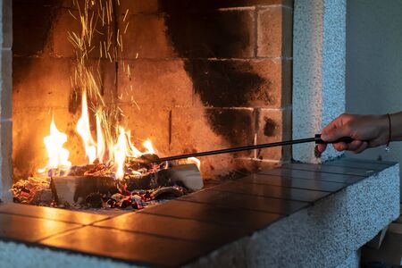 Female hand straightens burning firewood with an iron poker in an open fireplace and from the coal fly beautiful spark. Warmth and comfort in a cozy house, in winter.の写真素材