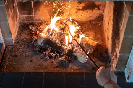 Female hand straightens and overturns burning firewood with an iron poker in an open fireplace and from the coal fly beautiful spark. Warmth and comfort in a cozy house, in the cold winter.の写真素材