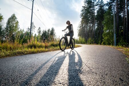 Happy woman rides a bike on a bicycle path near a pine forest. She is smiling and enjoying warm summer rain and the sun.の写真素材