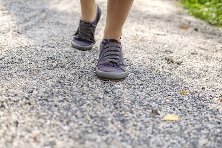 Female legs in sneakers rise up the trail of gravel on a sunny summer day, on a blurred background....
