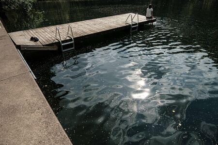 Woman sits on edge of a wooden pier with legs dipped in calm water of a clean, shiny forest lake, she dreams and enjoys the rays of morning sun.の写真素材