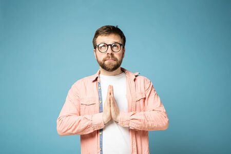 Calm, bearded man with glasses, cupped palms together and looks up. Isolated on a blue background.の写真素材