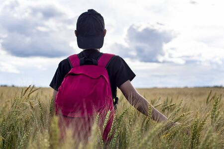Pensive, dreaming teenager with a backpack stands on a wheat field triticum and touches the ear with hands, against the sky and clouds. Vacation. Concept of freedom and relaxation.の写真素材