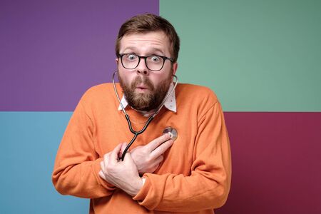 Surprised bearded man listens to his heart with a stethoscope and fearfully looks into the camera with big eyes.の写真素材