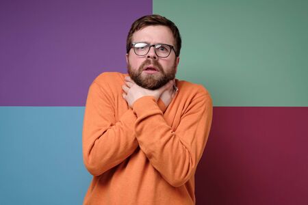Bearded man suffocates himself or feels a sore throat, holding neck with his hands, and looks frightened at the camera.の写真素材