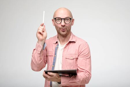 Brilliant idea. Intelligent man with glasses, indicates stylus pen up, holds a tablet in hands, looks joyfully at the camera.の写真素材