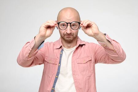 Bald, unshaven man took off glasses, keeping them at a distance, he examines the cleanliness of the lenses and looks funny.の写真素材