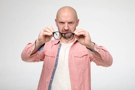 Upset bald man holds glasses in hands and examines them with alarm, he sees marriage on the lenses and is very angry.の写真素材