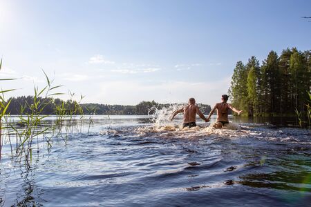 Man and a woman hold hands, swim and have fun in a clear forest lake on a hot summer day against a background of trees and sky. Active tourist life.の写真素材