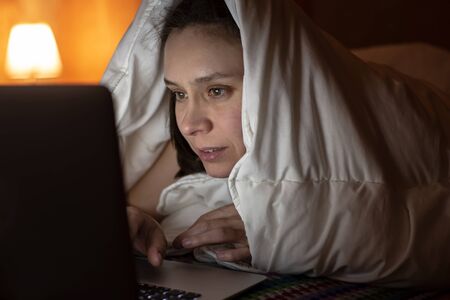 Woman looks with interest at a laptop, hiding under a blanket, in bed, at night by the light of a...