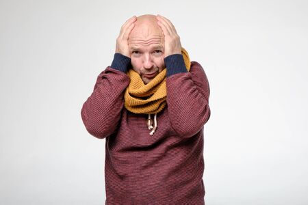 Man in a sweatshirt and scarf, trying to hide and warm his freezing, bald head, he covers it with hands.の写真素材