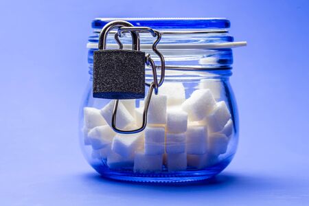 Cubes of white sugar in a transparent glass jar with a lid closed on a lock, on a blue background. Concept of proper nutrition. Close-up.の写真素材