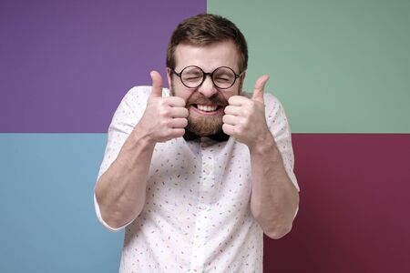 Funny bearded man in round glasses, makes an approving gesture with hands, thumbs up, closes eyes and grins teeth.の写真素材