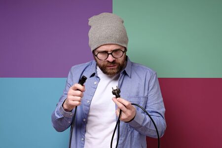 Puzzled bearded man, in round glasses and a hat, with a serious look looks at the plug with a wire, which he holds in hands.の写真素材