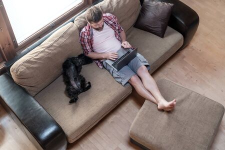 Young man works with a laptop, he is sitting on a soft sofa, next to him of his dog Schnauzer is sleeping with stomach upstairs, in a cozy home environment. Top view.の写真素材