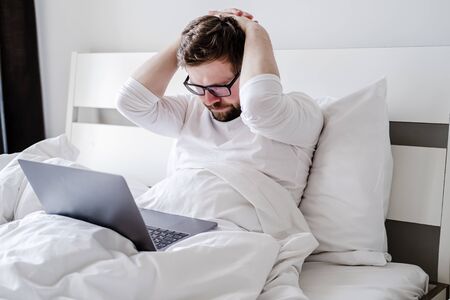 Man in pajamas is upset, he worked all night with a laptop in bed and did not finish the project, he holds hands behind head and looks thoughtfully at the screen.の写真素材