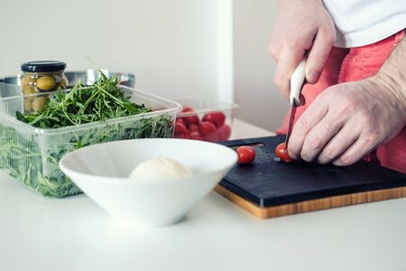 Man cuts tomatoes on a cutting board, on the table, next to arugula, olives and mozzarella in a plate. Concept of healthy food and home cooking. Close-up.の写真素材