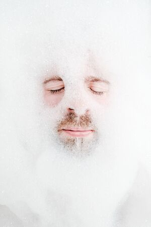 Male face in a lush foam, only eyes and mouth are visible, close-up. Man enjoys and relaxes while lying in a bathtub, with soapy water. Top view.の写真素材
